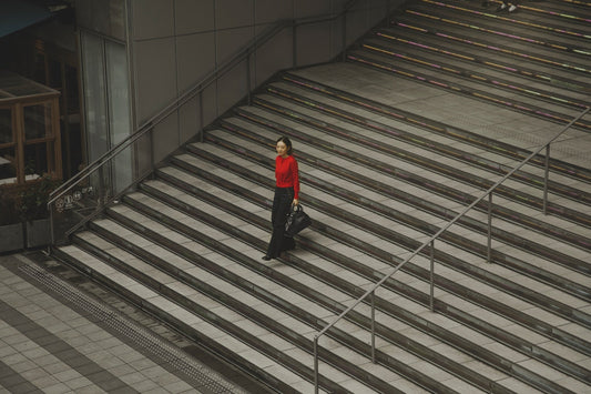 Hisae Takahashi in a red top and black denim, carrying the TSUCHIYA KABAN Clarte Arc Carry Tote in Black while walking up urban stairs.