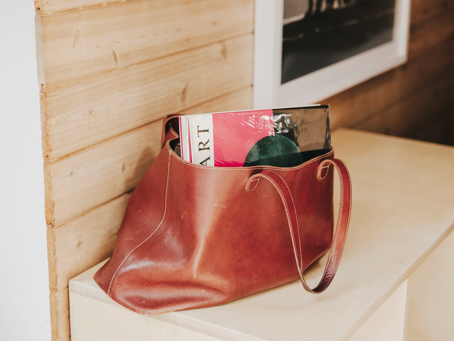 Brown leather tote bag with a book inside on a wooden surface.