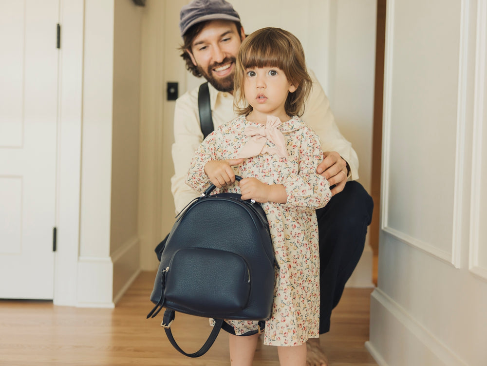 Small daughter playing with a backpack while her father watches