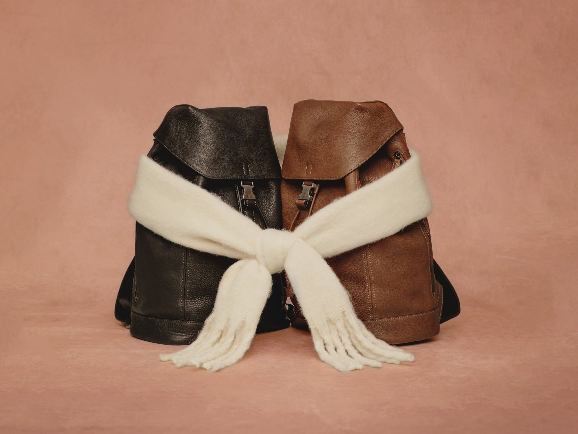 Holiday Collection: Two leather backpacks, one black and one brown, with a white scarf draped over them on a pink background. 