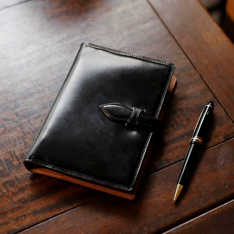 Black leather planner with a decorative clasp on a wooden surface next to a pen.