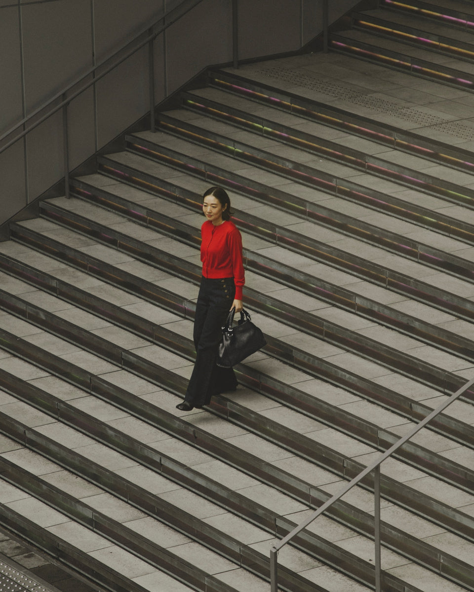 Hisae Takahashi in a red top and black denim, carrying the TSUCHIYA KABAN Clarte Arc Carry Tote in Black while walking up urban stairs.