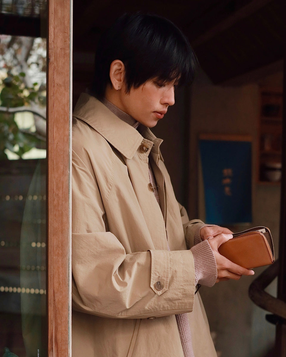 Person in a beige trench coat holding a brown leather Cordovan Long wallet 