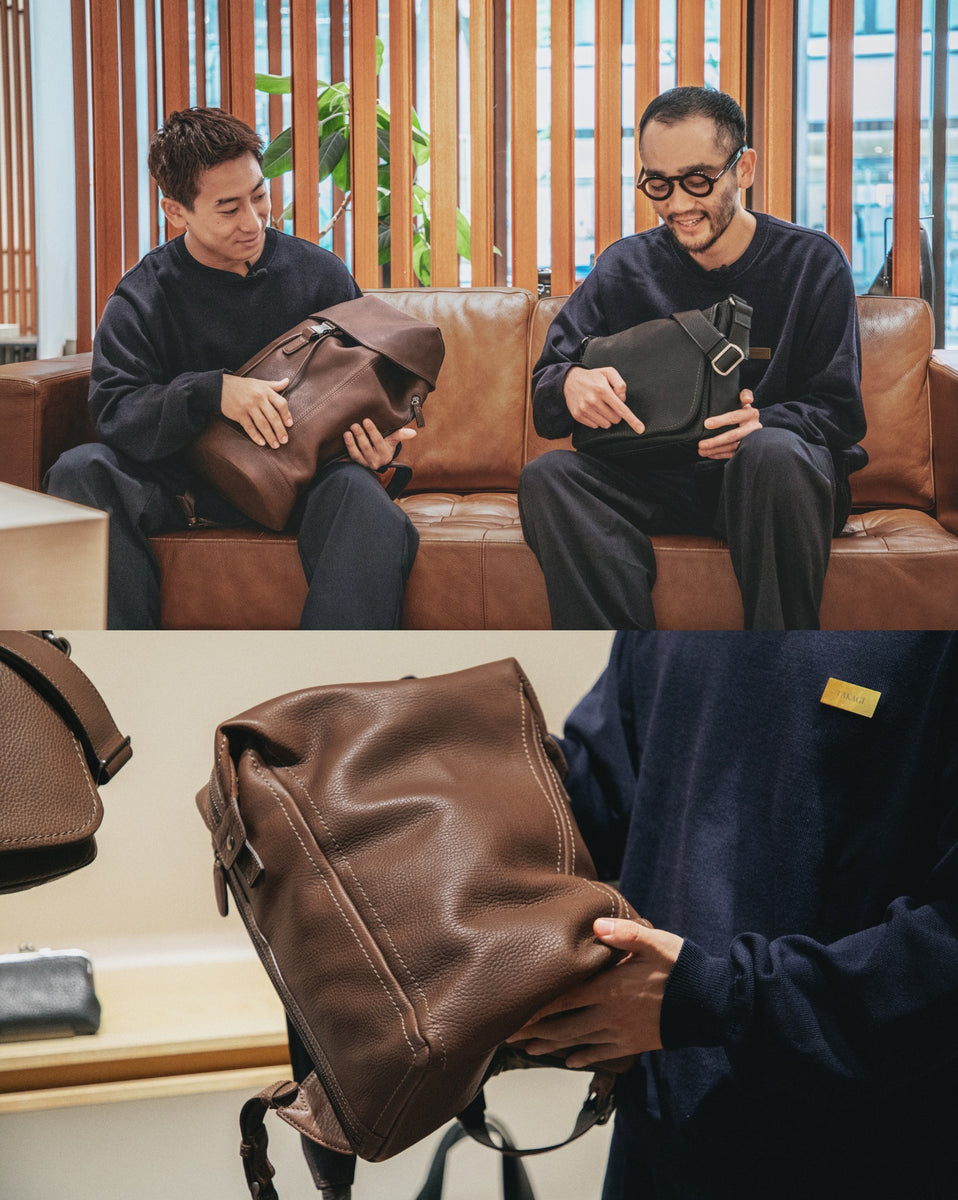 Two Tsuchiya staff sitting on a brown leather couch in a modern indoor setting, each holding a leather bag from Tsuchiya Kaban's Tone Nume collection. The man on the left holds a brown leather bag, and the man on the right holds a black leather bag with a silver buckle.