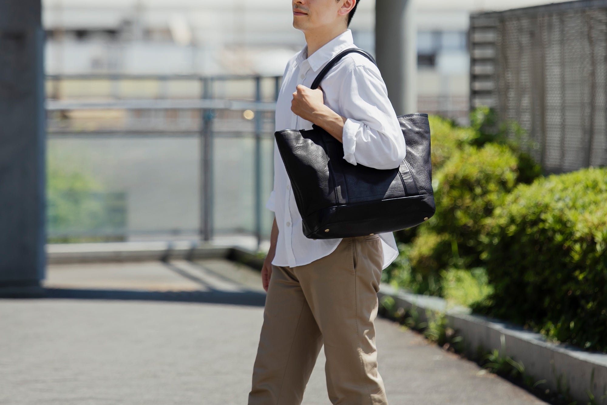 A man in a white button-down shirt and khaki pants carrying a Dark Indigo Roadster Large leather tote bag over his shoulder, walking outdoors on a sunny day.