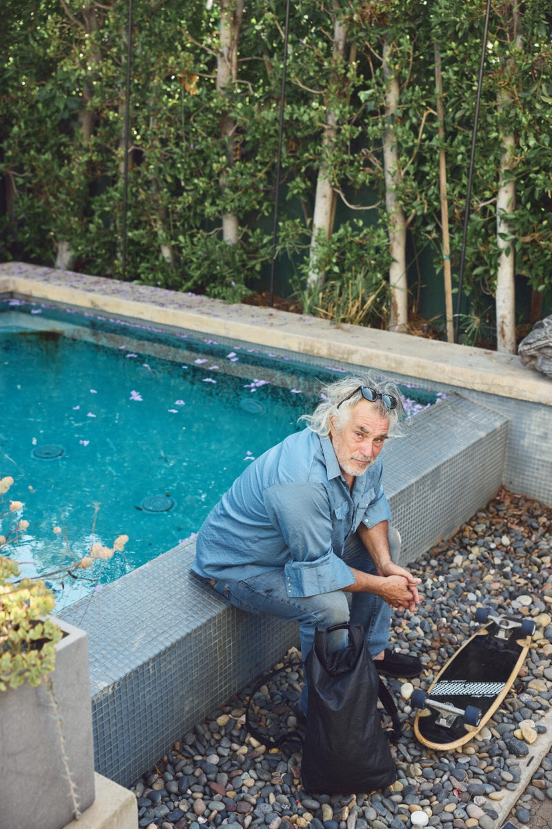 Steve Olson sits on a tiled poolside ledge wearing a light blue denim shirt and jeans, with a black Tsuchiya Kaban Rolltop backpack on the ground beside him and a skateboard nearby. The setting features a swimming pool, gravel, plants, and a stone wall in the background.