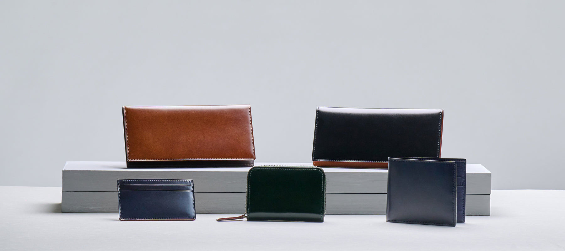 The image shows a collection of five Tsuchiya Kaban's leather wallets of various sizes and colors arranged on a white and gray display. 