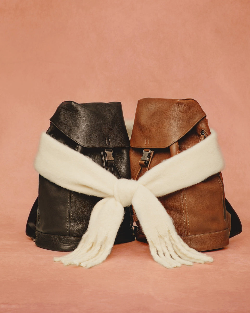 Holiday Collection: Two leather backpacks, one black and one brown, with a white scarf draped over them on a pink background. 