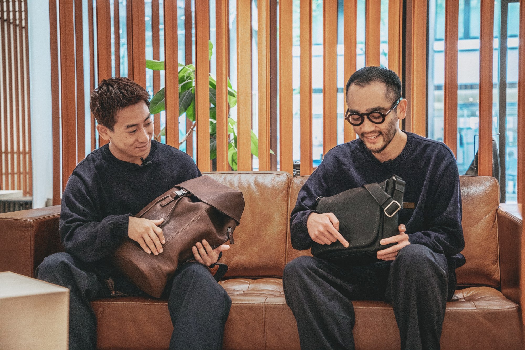 Two Tsuchiya staff sitting on a brown leather couch in a modern indoor setting, each holding a leather bag from Tsuchiya Kaban's Tone Nume collection. The man on the left holds a brown leather bag, and the man on the right holds a black leather bag with a silver buckle.