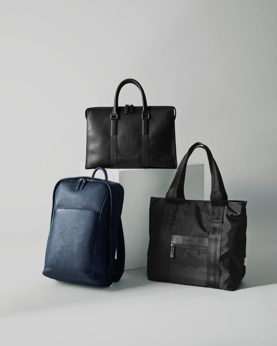 Three bags displayed on a light gray studio background: a black leather briefcase-style top bag on a pedestal, a navy leather backpack, and a black nylon tote bag with handles and a front zip pocket.