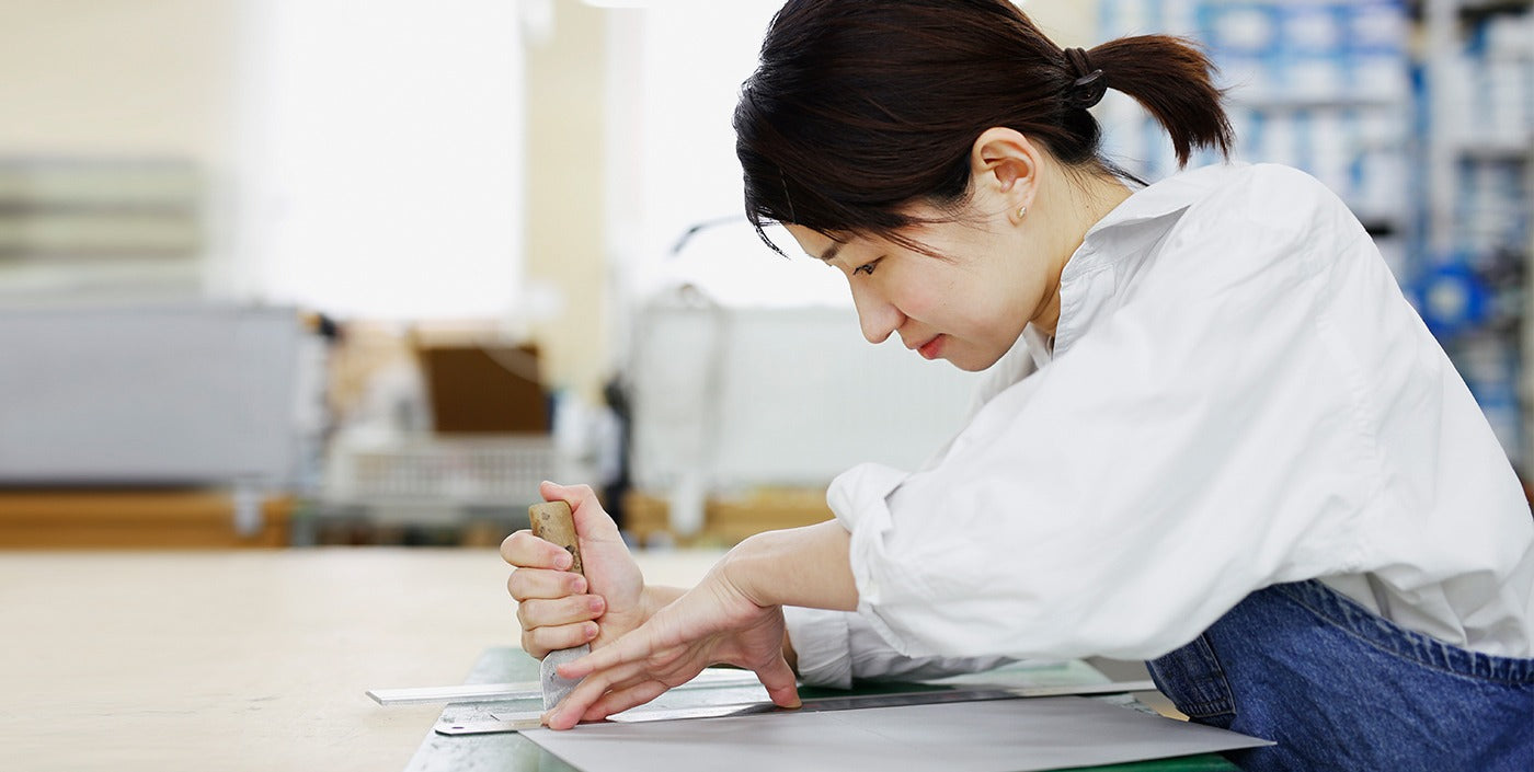 Ms. Sasaki, Tsuchiya Kaban's craftsperson, focused on cutting material with a craft knife and a ruler on a workbench.