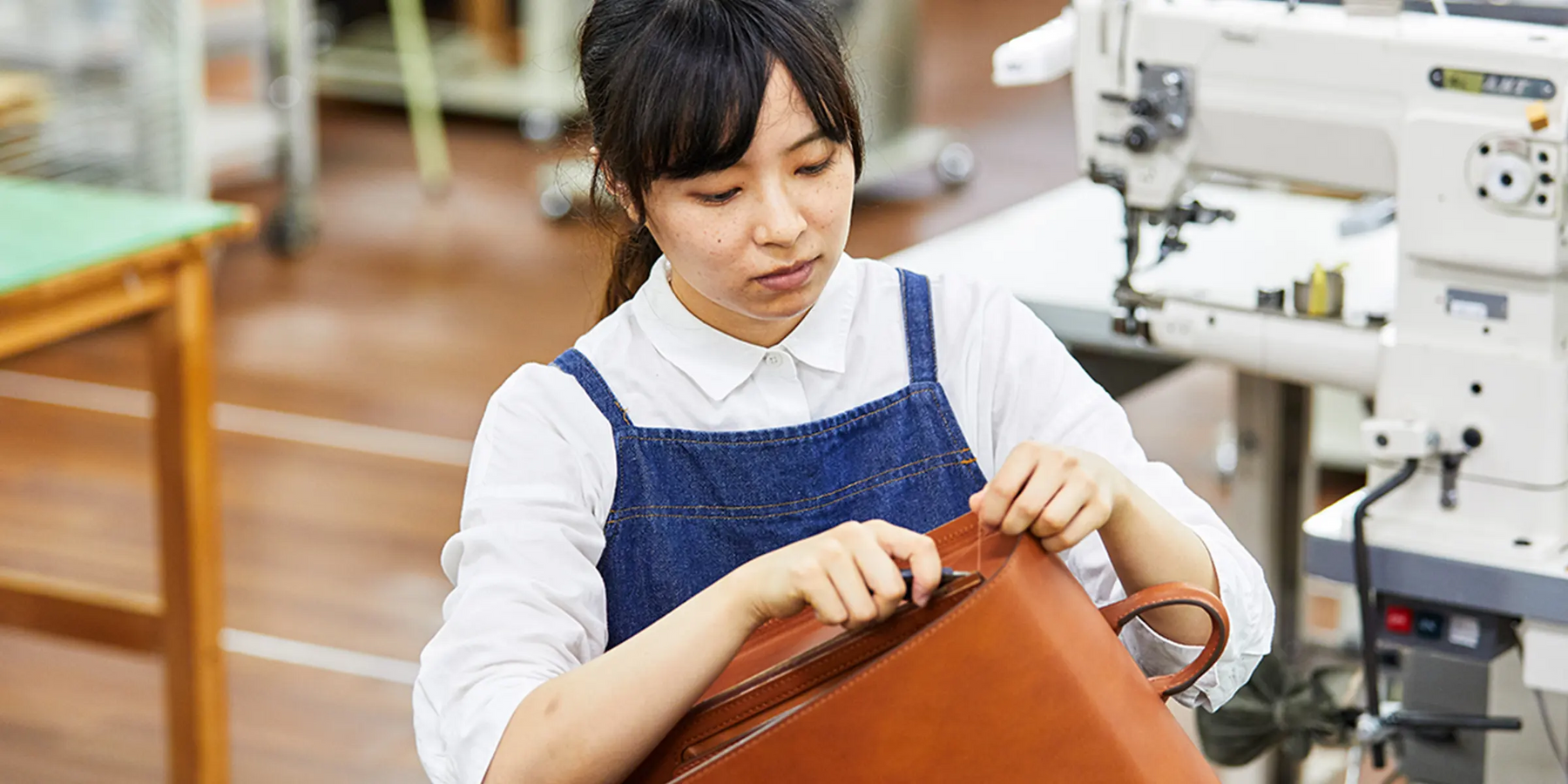 Eri Sugita, a craftsperson at Tsuchiya Kaban, is depicted in a workshop setting, deeply engrossed in working on a brown leather bag. She is wearing a white collared shirt under a denim apron, which is her typical work attire.