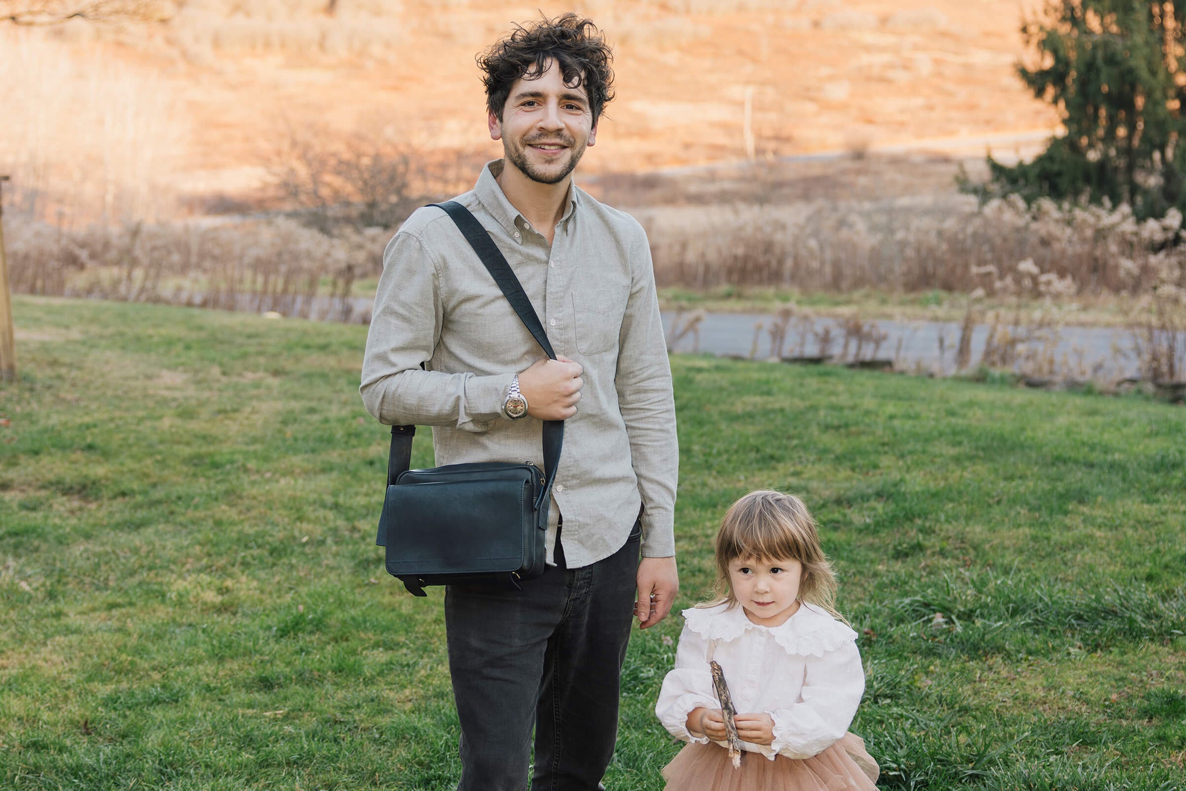 Father with a dark navy Roadster leather crossbody bag standing next to a young girl, his daughter in a dress on the grass