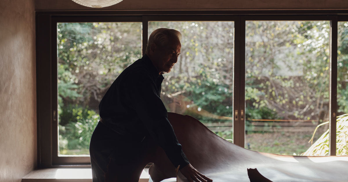 Kunio Tsuchiya of Founder of Tsuchiya Kaban, seen in profile, stands in a room with large windows that look out onto a lush green, sunlit garden. He is dressed in a dark, long-sleeved shirt and appears to be smoothing or adjusting a large piece of material, possibly leather or fabric, laid out on a surface in front of him. The room has a warm, natural light entering from the windows, which also reflect some of the interior.